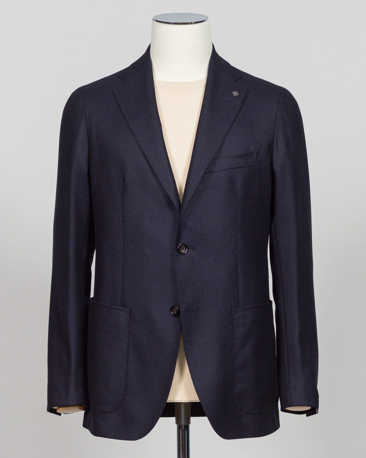 A dark navy blue blazer displayed on a white mannequin torso, layered over a light beige shirt, against a neutral gray background. Italian wool-cashmere sportcoat engineered for refined elegance. The blend softens with wear while maintaining structural integrity. Navy's warmth anchors tailored trousers and raw denim alike. Structured shoulders, fluid drape, precise construction—details that reward inspection. A jacket for intentional dressing, equally suited to client meetings, dinners, or elevated casual w