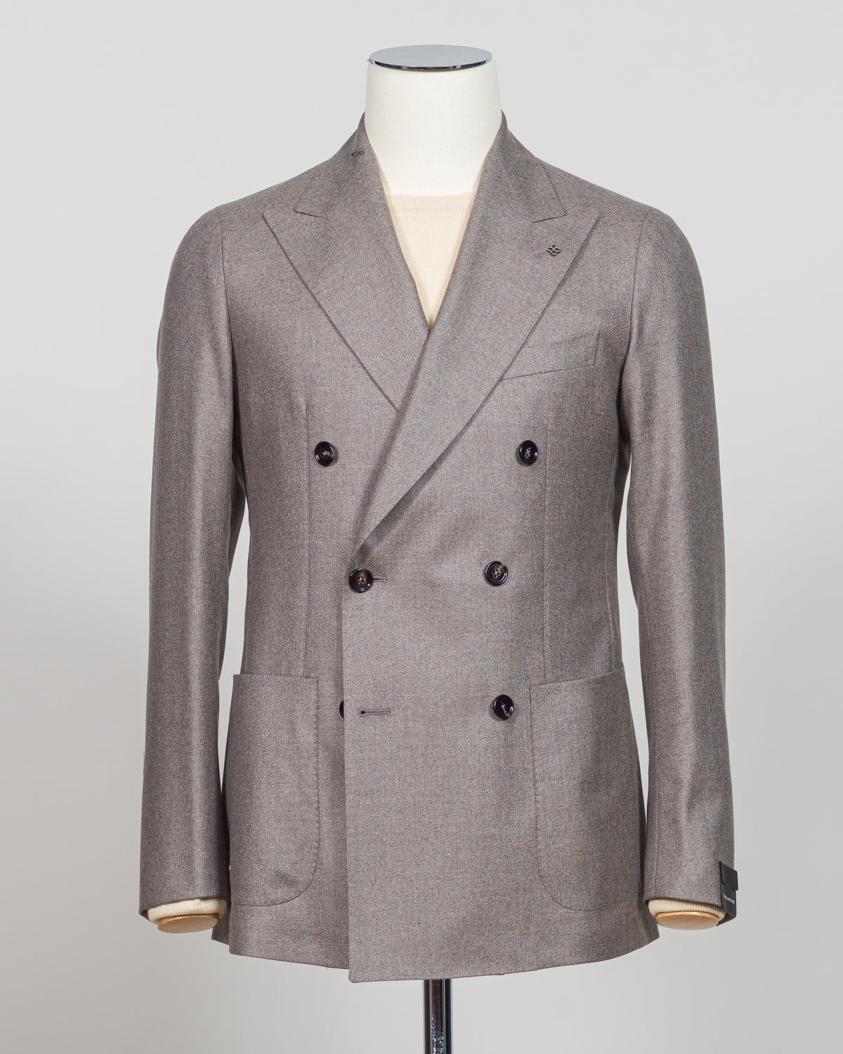 A double-breasted blazer in a textured taupe-gray fabric, displayed on a white mannequin torso. The blazer features a wrap-style collar, six dark buttons, and front patch pockets, with a light beige shirt visible underneath. Tagliatore Double-Breasted Jacket / Taupe A refined double-breasted silhouette from Tagliatore, crafted in a sophisticated taupe that bridges season and wardrobe. The structured shoulders and clean lines define a timeless sport jacket suited to both tailored and relaxed styling. Italian