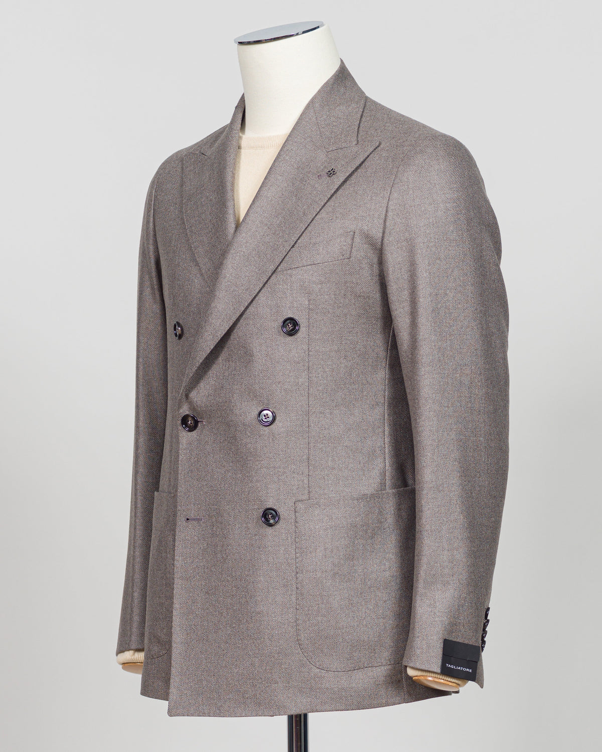 A double-breasted blazer in a textured taupe-gray fabric, displayed on a cream dress form mannequin against a light gray background. Tagliatore Double-Breasted Jacket / Taupe A refined double-breasted silhouette from Tagliatore, crafted in a sophisticated taupe that bridges season and wardrobe. The structured shoulders and clean lines define a timeless sport jacket suited to both tailored and relaxed styling. Italian construction meets versatile neutrality—a foundational piece for the discerning wardrobe