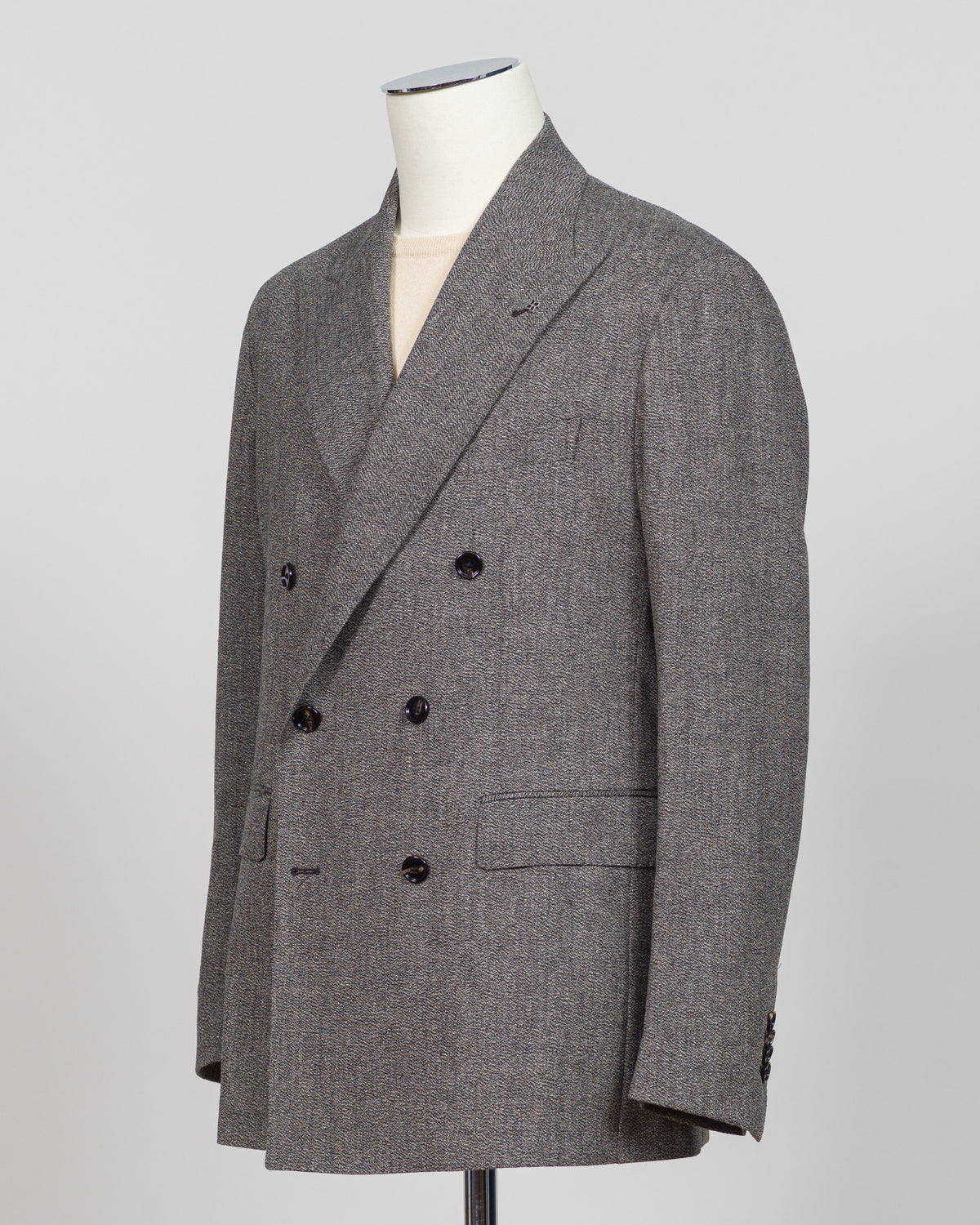 A gray double-breasted blazer with a subtle herringbone pattern, displayed on a white mannequin torso against a light gray background. Tagliatore's double-breasted silhouette merges Italian craftsmanship with refined restraint. The brown and grey palette offers versatility across seasons and occasions—from boardroom formality to sophisticated evening wear. Carefully finished details and structured shoulders create a commanding presence while maintaining contemporary proportion. Cut for the discerning wearer