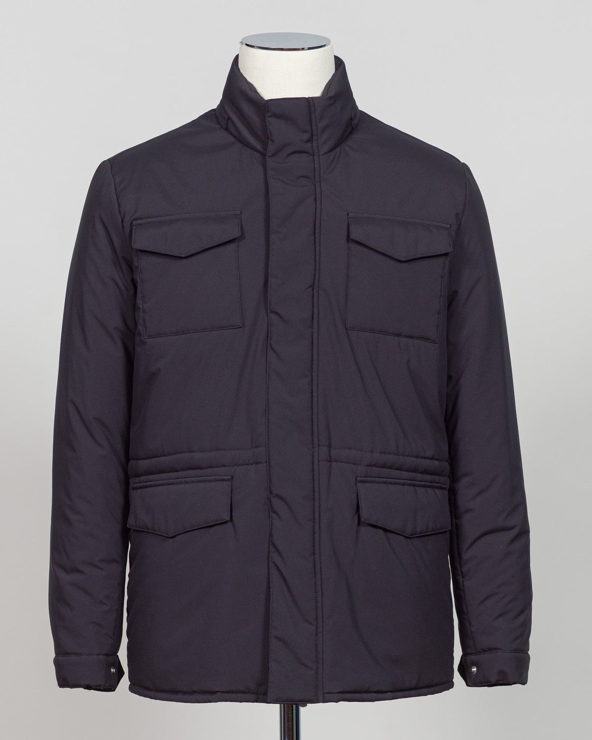 A dark navy blue quilted jacket displayed on a white mannequin torso against a neutral light gray The Gran Sasso Padded Nylon Jacket in navy delivers genuine warmth without the weight. Its lightweight thermowadding construction and refined collar offer understated sophistication—equally at home over a dress shirt or layered with knitwear. Italian craftsmanship meets everyday practicality: polished for client meetings, reliable for daily wear, and versatile enough to become your most trusted layerbackground.