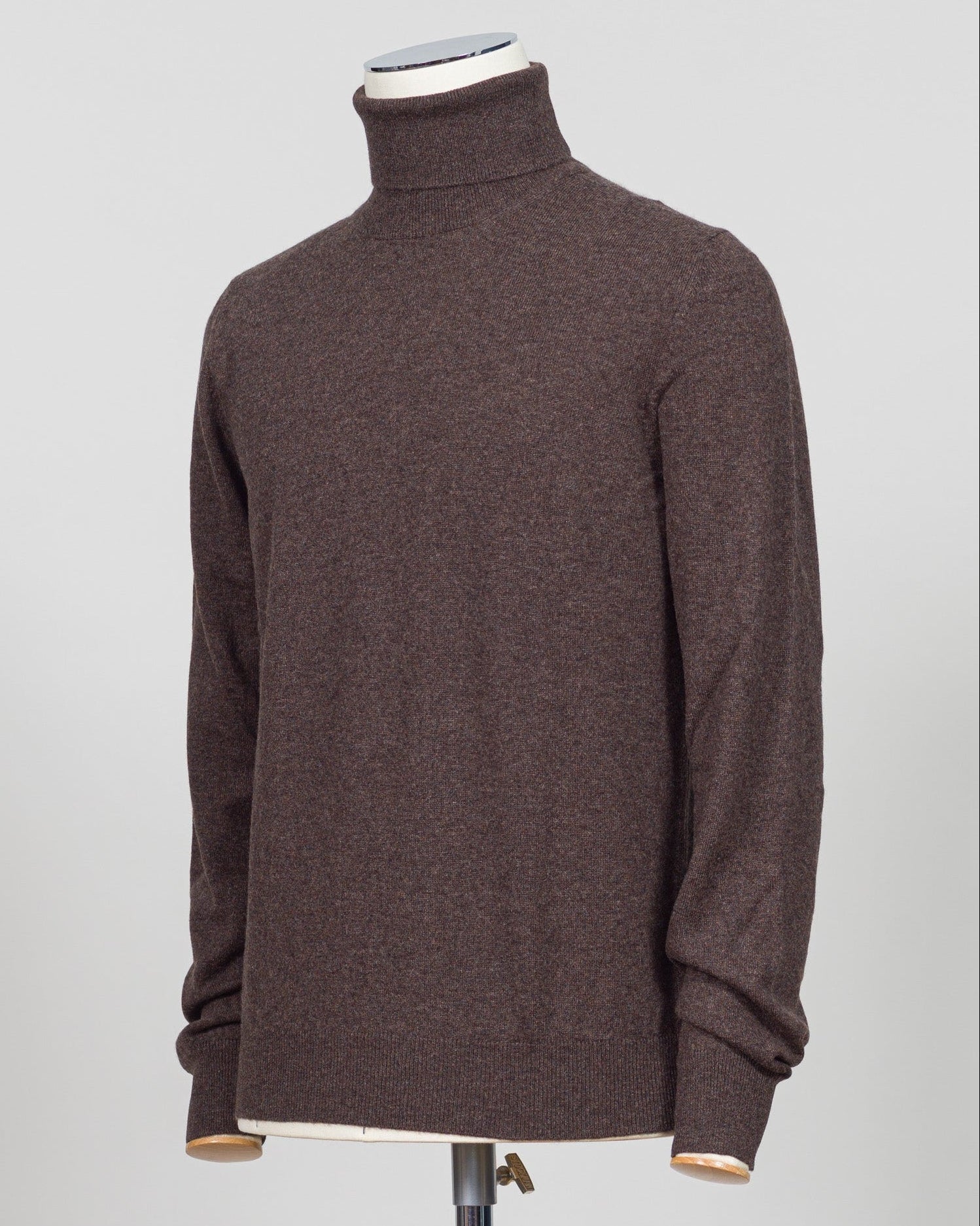 A dark brown knit turtleneck sweater displayed on a mannequin torso against a light gray studio background. Italian craftsmanship meets understated elegance. This Gran Sasso cashmere roll neck in oat delivers refined warmth through premium fibers and meticulous construction. A versatile layering piece that anchors both formal and relaxed wardrobes with timeless appeal. Gran Sasso Cashmere Roll Neck / Dark Brown