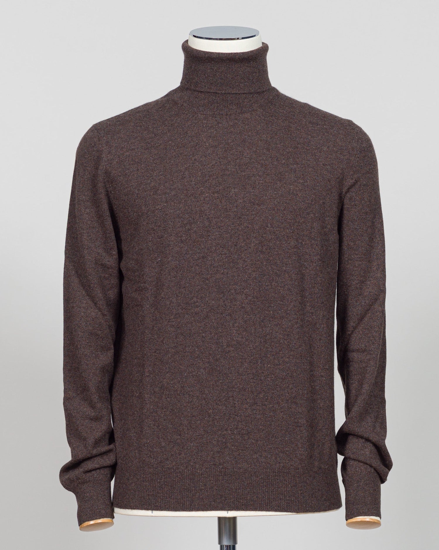 A dark brown knit turtleneck sweater displayed on a mannequin torso against a light gray background. Italian craftsmanship meets understated elegance. This Gran Sasso cashmere roll neck in oat delivers refined warmth through premium fibers and meticulous construction. A versatile layering piece that anchors both formal and relaxed wardrobes with timeless appeal. Gran Sasso Cashmere Roll Neck / Dark Brown