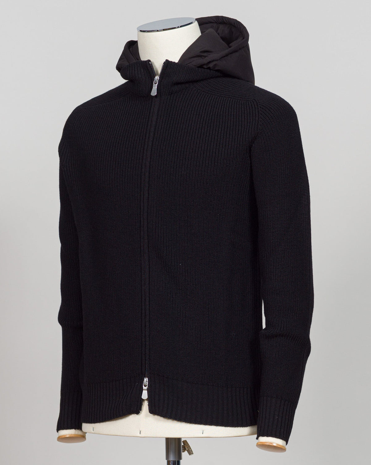A black ribbed knit zip-up hoodie with a hood, displayed on a cream-colored mannequin torso against a light gray background. Italian craftsmanship meets weatherproof functionality in this Gran Sasso rain wool hoodie. Constructed from premium wool blended with water-resistant properties, it delivers warmth and protection without bulk. The relaxed fit and classic black colorway make it a versatile layering essential for unpredictable Nordic weather. Finished with refined detailing befitting Gran Sasso's herit