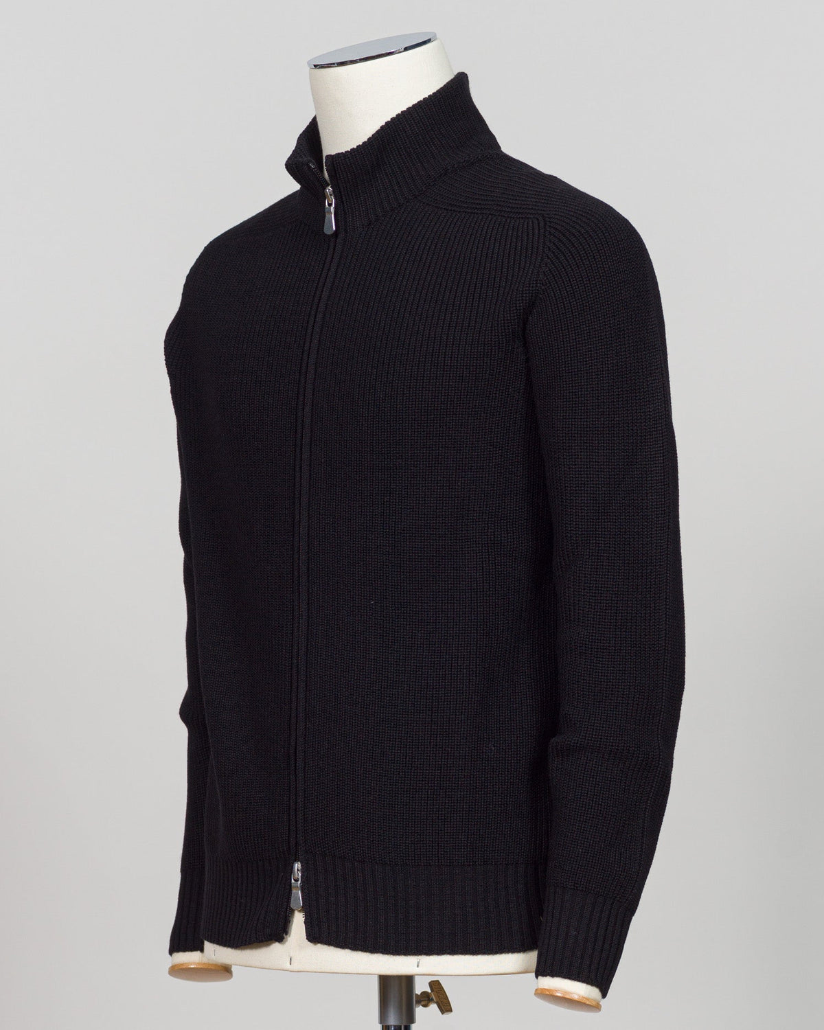 A black ribbed knit zip-up sweater displayed on a cream mannequin torso against a light gray studio background. Italian craftsmanship meets weatherproof functionality in this Gran Sasso rain wool hoodie. Constructed from premium wool blended with water-resistant properties, it delivers warmth and protection without bulk. The relaxed fit and classic black colorway make it a versatile layering essential for unpredictable Nordic weather. Finished with refined detailing befitting Gran Sasso's heritage of fine k