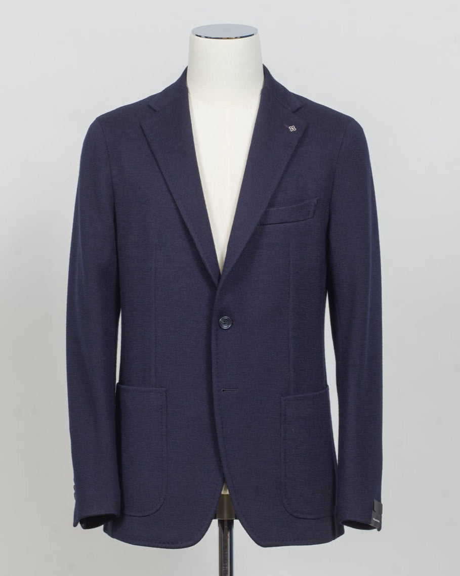 A navy blue blazer displayed on a white mannequin torso against a light gray studio background. Tailored jersey jacket in soft navy wool blend. Clean, modern silhouette designed for everyday sophistication. Lightweight construction offers comfort without sacrificing structure. Perfect for layering or wearing solo across seasons. Italian craftsmanship meets versatile styling. Tagliatore Jersey Jacket / Navy