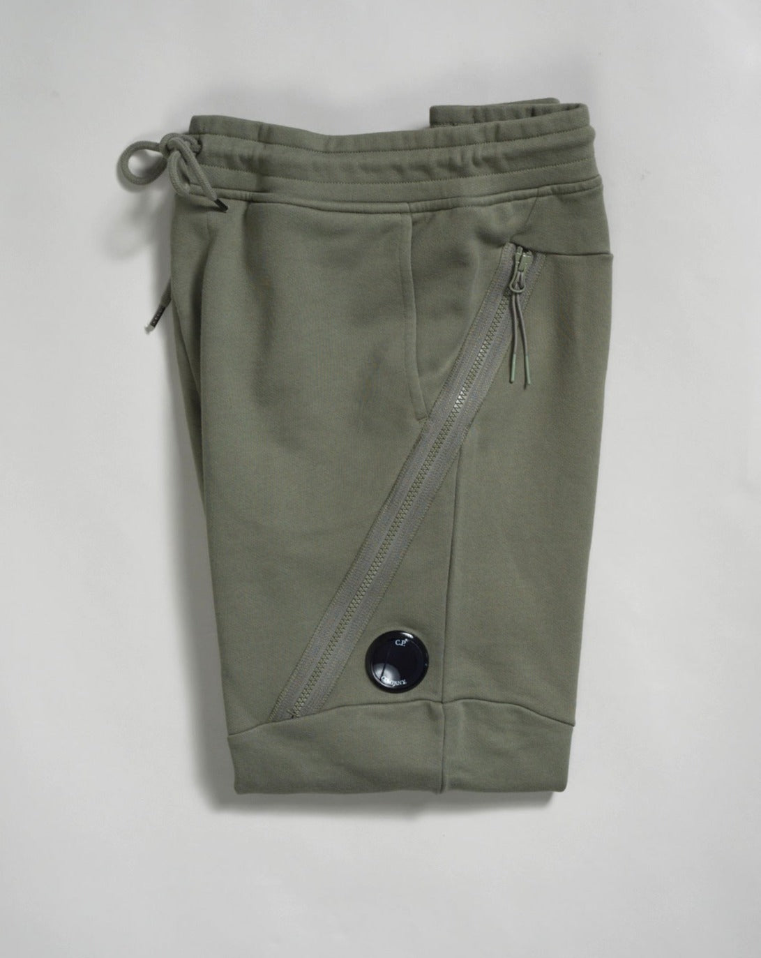 Sweatpants with an adjustable drawstring waistband and tapered legs. The sweatpants also feature slanted hand pockets and a C.P. Company Lens detail on the left leg. Crafted in diagonal raised fleece, a midweight loopback cotton option that combines absolute comfort with a substantial and hard-wearing feel. 14CMSP084A005086W Col 648 Bronze Green Adjustable drawstring waistband Slanted hand pockets Angular thigh zip pockets Lens detail Tapered leg
