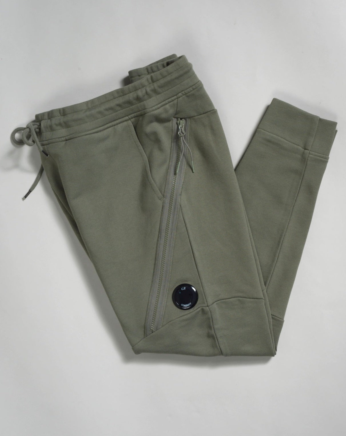 Sweatpants with an adjustable drawstring waistband and tapered legs. The sweatpants also feature slanted hand pockets and a C.P. Company Lens detail on the left leg. Crafted in diagonal raised fleece, a midweight loopback cotton option that combines absolute comfort with a substantial and hard-wearing feel. 14CMSP084A005086W Col 648 Bronze Green Adjustable drawstring waistband Slanted hand pockets Angular thigh zip pockets Lens detail Tapered leg