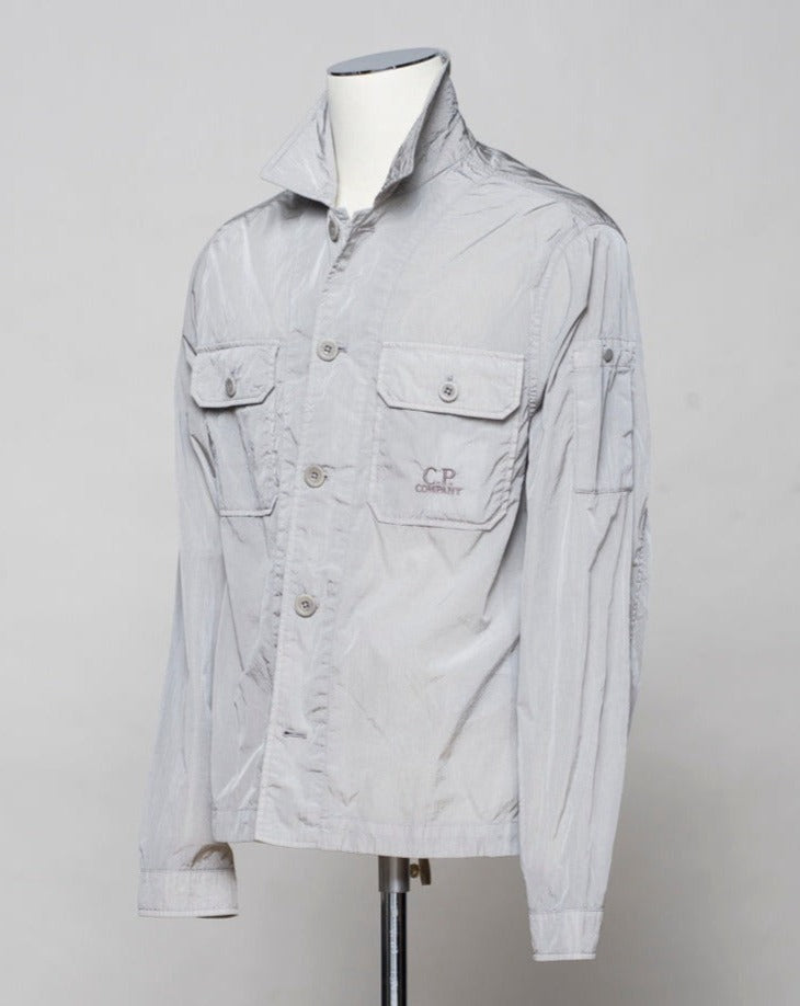 Button fastening in front. 2 breast pockets with buttons. Small pocket on left arm with snap fastener. Embroidered tone-on-tone C.P. Company logo on left pocket. Article: 16CMOS106A 005904G. Composition: 100% pre-consumer recycled nylon. Garment dyed. Color: 913 / Drizzle Grey