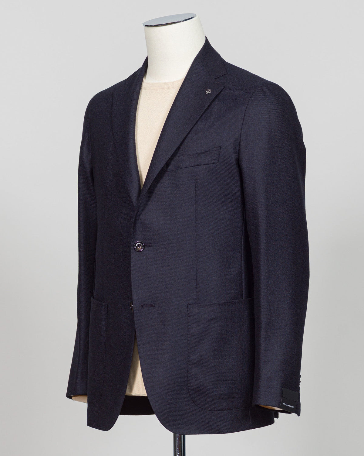 A dark navy blue blazer displayed on a mannequin torso over a light beige shirt, set against a plain light gray background. Italian wool-cashmere sportcoat engineered for refined elegance. The blend softens with wear while maintaining structural integrity. Navy's warmth anchors tailored trousers and raw denim alike. Structured shoulders, fluid drape, precise construction—details that reward inspection. A jacket for intentional dressing, equally suited to client meetings, dinners, or elevated casual wear. Ta