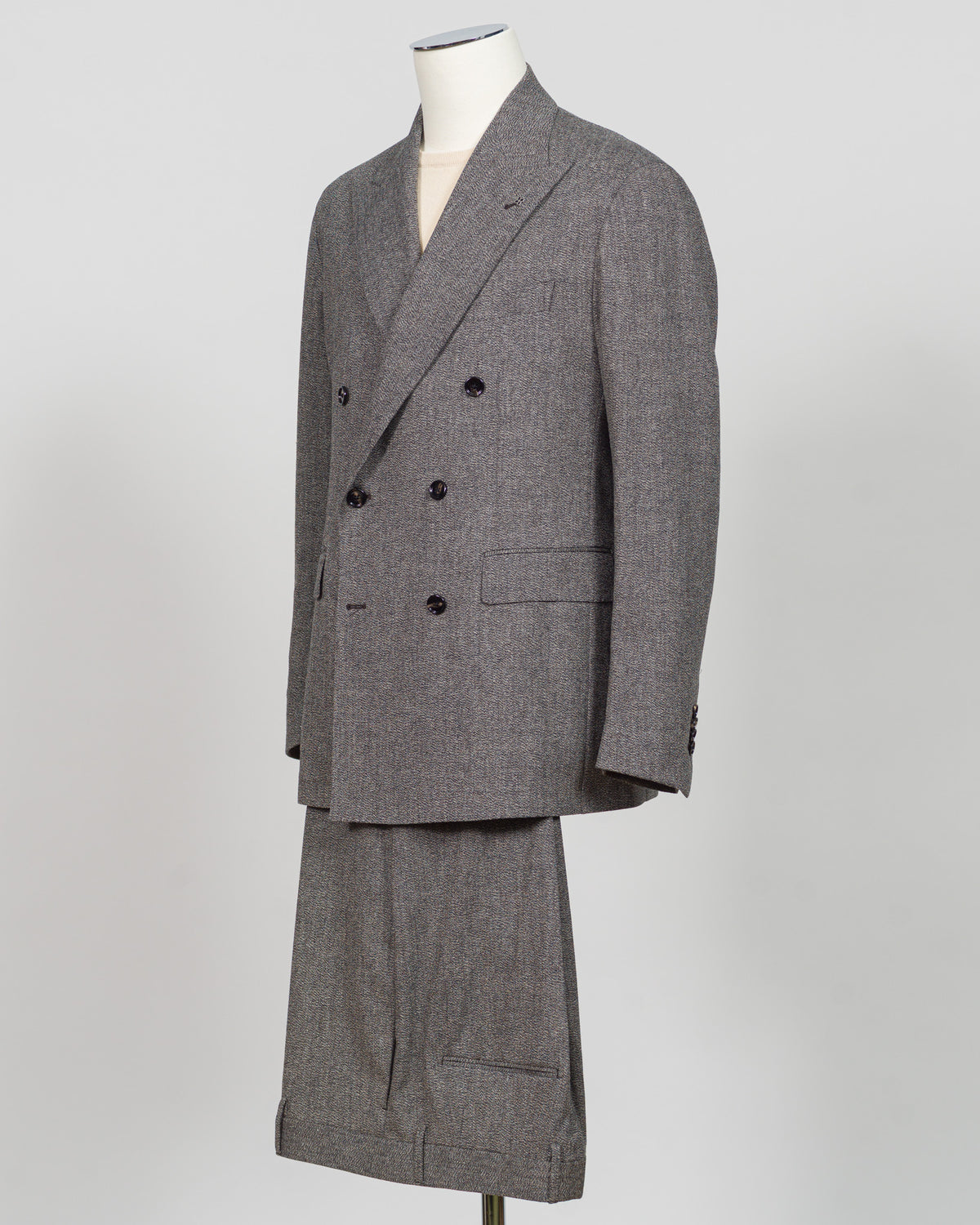 A gray double-breasted suit with a subtle houndstooth pattern, displayed on a mannequin against a light gray background. Tagliatore's double-breasted silhouette merges Italian craftsmanship with refined restraint. The brown and grey palette offers versatility across seasons and occasions—from boardroom formality to sophisticated evening wear. Carefully finished details and structured shoulders create a commanding presence while maintaining contemporary proportion. Cut for the discerning wearer who values ti