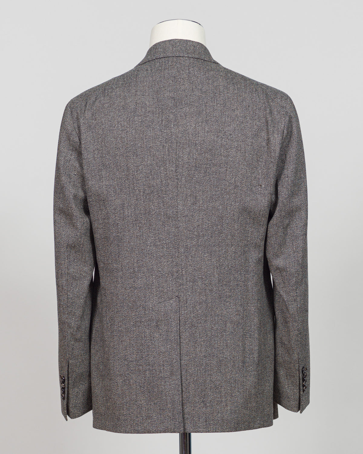 The back view of a men's gray herringbone tweed blazer displayed on a white mannequin torso against a light gray background. Tagliatore's double-breasted silhouette merges Italian craftsmanship with refined restraint. The brown and grey palette offers versatility across seasons and occasions—from boardroom formality to sophisticated evening wear. Carefully finished details and structured shoulders create a commanding presence while maintaining contemporary proportion. Cut for the discerning wearer who value