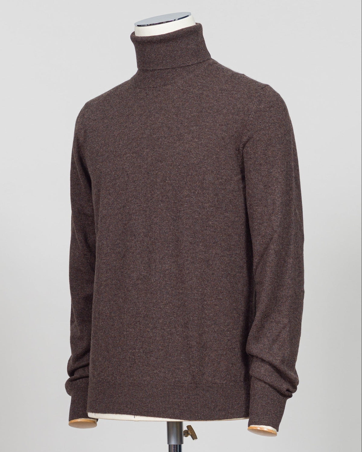 A dark brown knit turtleneck sweater displayed on a mannequin torso against a light gray studio background. Italian craftsmanship meets understated elegance. This Gran Sasso cashmere roll neck in oat delivers refined warmth through premium fibers and meticulous construction. A versatile layering piece that anchors both formal and relaxed wardrobes with timeless appeal. Gran Sasso Cashmere Roll Neck / Dark Brown
