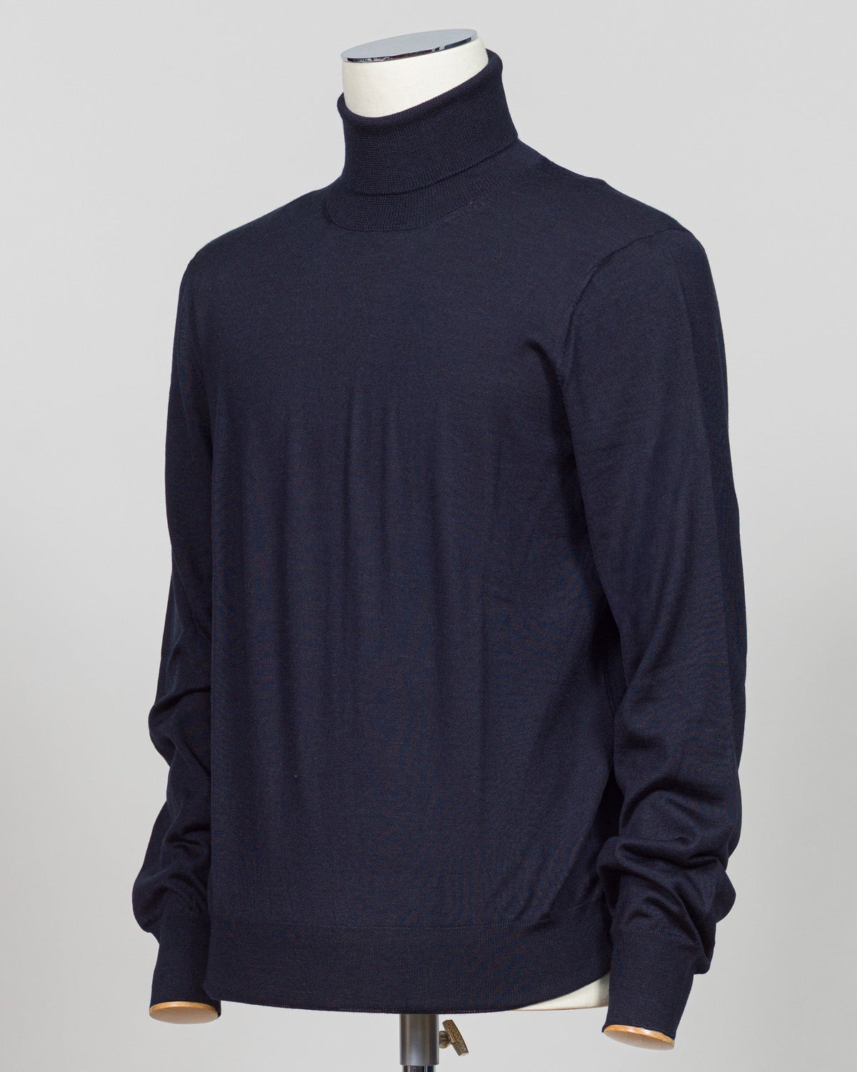 A dark navy blue knit turtleneck sweater displayed on a white mannequin torso against a light gray  Refined luxury knitwear from Gran Sasso, combining wool, cashmere, and silk in a timeless roll neck silhouette. The navy colorway offers versatile sophistication—equally at home layered under tailoring or worn as a standalone piece. Soft, breathable construction ensures comfort across seasons, while the blend of natural fibers delivers lasting durability and subtle sheen.A wardrobe essentia studio background.