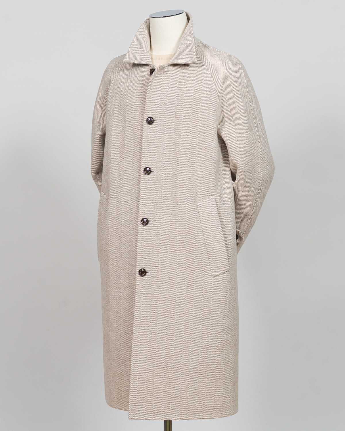 A light beige wool-blend coat displayed on a mannequin against a neutral light gray background. The coat features a classic collar, a front button closure with dark buttons, and large front patch pockets.