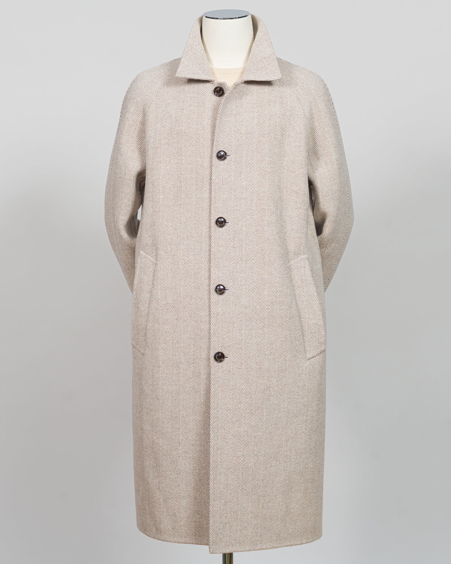 A light beige wool-blend coat displayed on a mannequin against a neutral light gray background. The coat features a classic collar, a front button closure with dark buttons, and large front patch pockets.