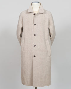 A light beige wool-blend coat displayed on a mannequin against a neutral light gray background. The coat features a classic collar, a front button closure with dark buttons, and large front patch pockets.
