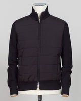 A black mixed-media jacket with a quilted body and ribbed knit sleeves, collar, and hem, displayed on a mannequin torso against a light gray background. Italian wool craftsmanship meets contemporary tailoring in this Gran Sasso sartorial padded jacket. Structured in premium wool with subtle padding for refined warmth, it pairs effortlessly with business wear or elevated casual styling. Gran Sasso Sartorial Padded &nbsp;Wool Jacket / Black