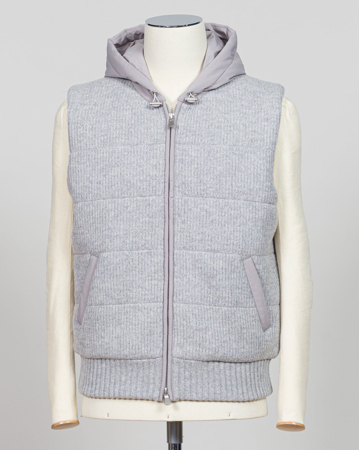 A light gray ribbed knit zip-up vest with a matching gray hood, displayed on a cream-colored mannequin against a light gray background. Lightweight insulation meets refined elegance in this Gran Sasso padded hooded vest. The soft light grey colourway pairs seamlessly with layered knitwear or worn solo over shirts for versatile warmth. Italian craftsmanship ensures a tailored silhouette that bridges casual and smart-casual dressing. Essential for the discerning wardrobe. Gran Sasso Padded Hooded Vest / Light
