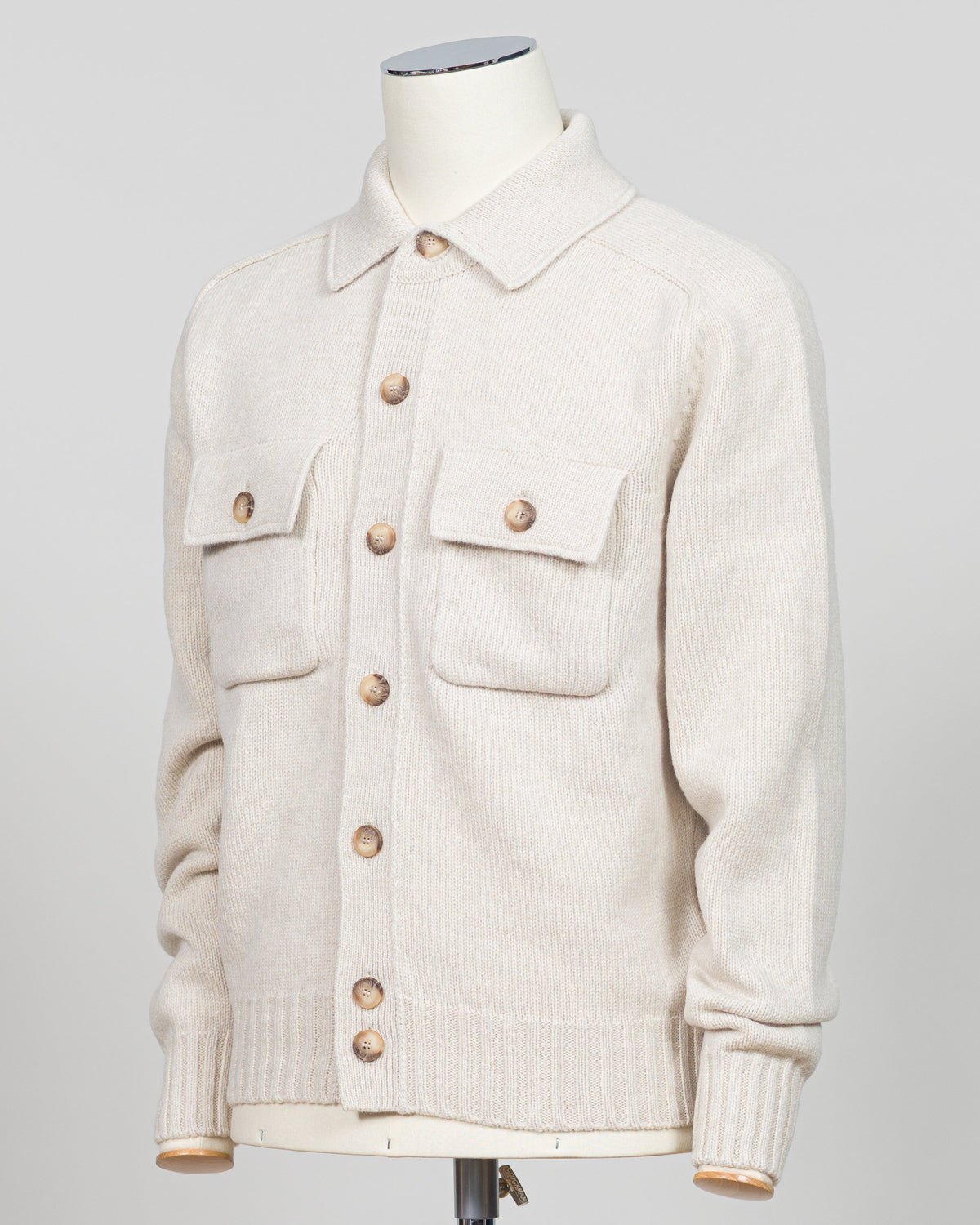 A cream-colored knit button-up cardigan sweater with flap chest pockets and wooden buttons, displayed on a mannequin torso against a light gray background.