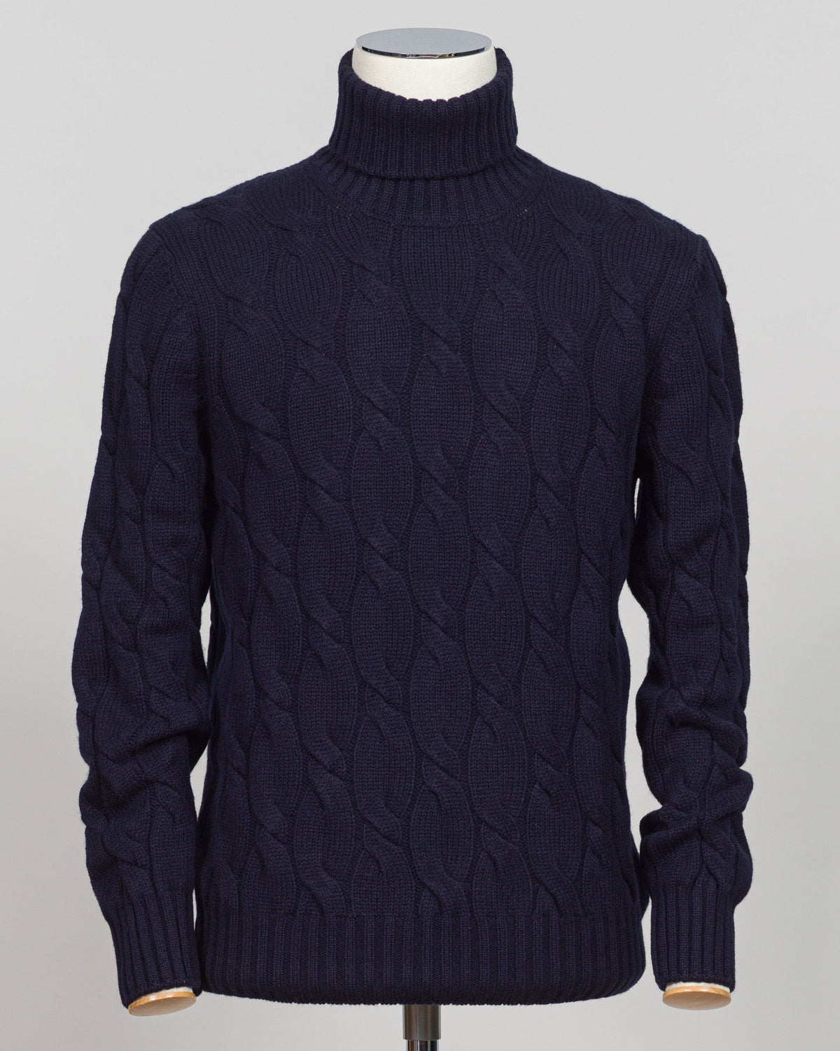 A dark navy blue cable-knit turtleneck sweater displayed on a mannequin torso against a light gray background. A timeless cable knit roll neck in deep navy. Crafted with refined construction, this piece layers effortlessly under tailored jackets or stands alone as an essential foundation for classic menswear. The textured cable pattern adds subtle depth while maintaining the understated elegance expected of premium knitwear. Gran Sasso Cable Roll Neck Knit / Navy