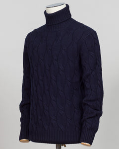 A dark navy blue cable-knit turtleneck sweater displayed on a mannequin torso against a light gray background. A timeless cable knit roll neck in deep navy. Crafted with refined construction, this piece layers effortlessly under tailored jackets or stands alone as an essential foundation for classic menswear. The textured cable pattern adds subtle depth while maintaining the understated elegance expected of premium knitwear. Gran Sasso Cable Roll Neck Knit / Navy