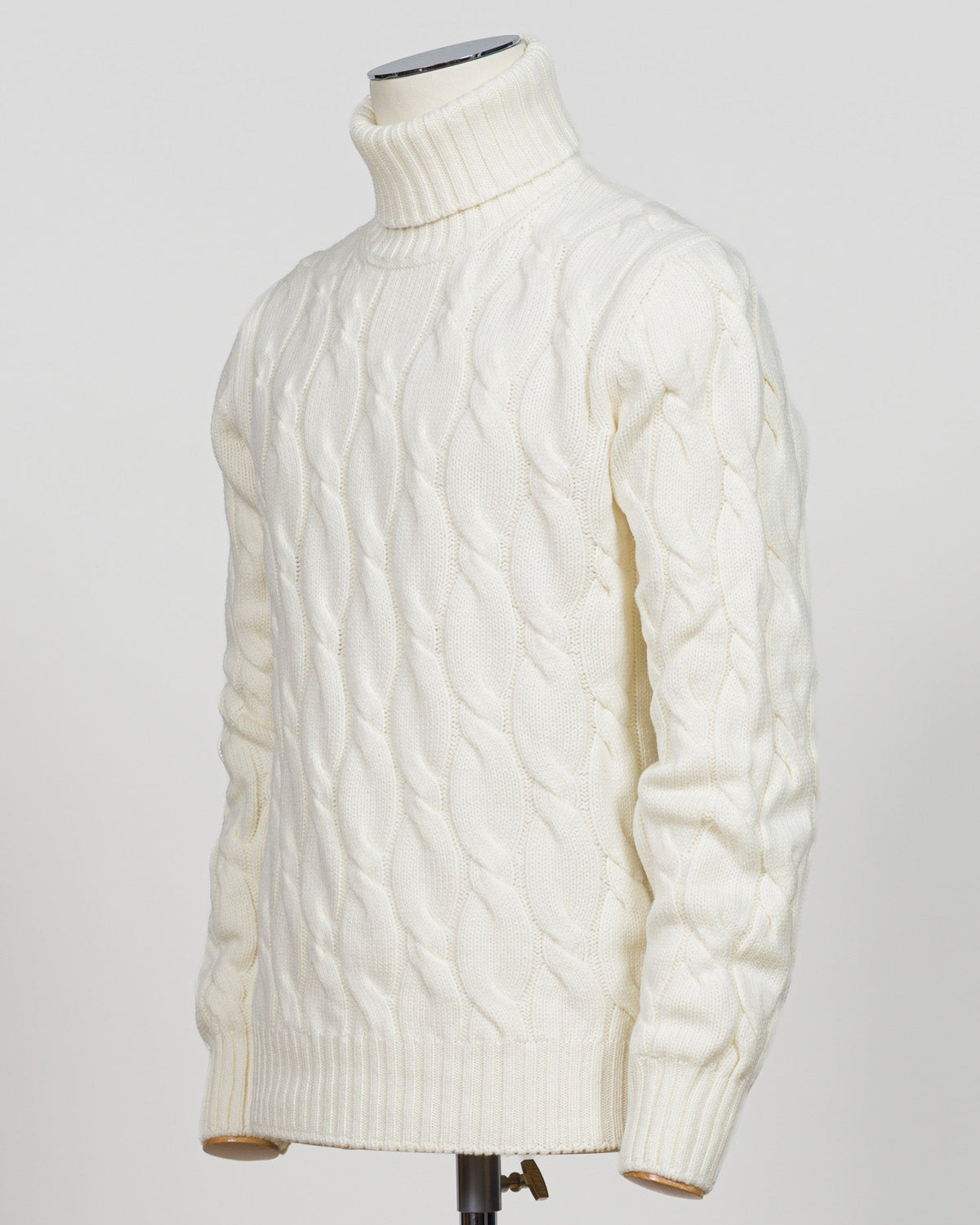 An off-white cable-knit turtleneck sweater displayed on a mannequin torso against a light gray background.  Italian craftsmanship meets timeless elegance in this Gran Sasso cable knit roll neck. Pure white wool construction delivers warmth and refined texture, while the classic silhouette transitions effortlessly from casual to smart-casual layering. A versatile essential for the considered wardrobe. Gran Sasso Cable Roll Neck Knit / White