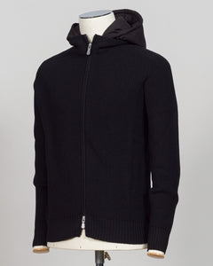 A black ribbed knit zip-up hoodie with a hood, displayed on a cream-colored mannequin torso against a light gray background. Italian craftsmanship meets weatherproof functionality in this Gran Sasso rain wool hoodie. Constructed from premium wool blended with water-resistant properties, it delivers warmth and protection without bulk. The relaxed fit and classic black colorway make it a versatile layering essential for unpredictable Nordic weather. Finished with refined detailing befitting Gran Sasso's herit