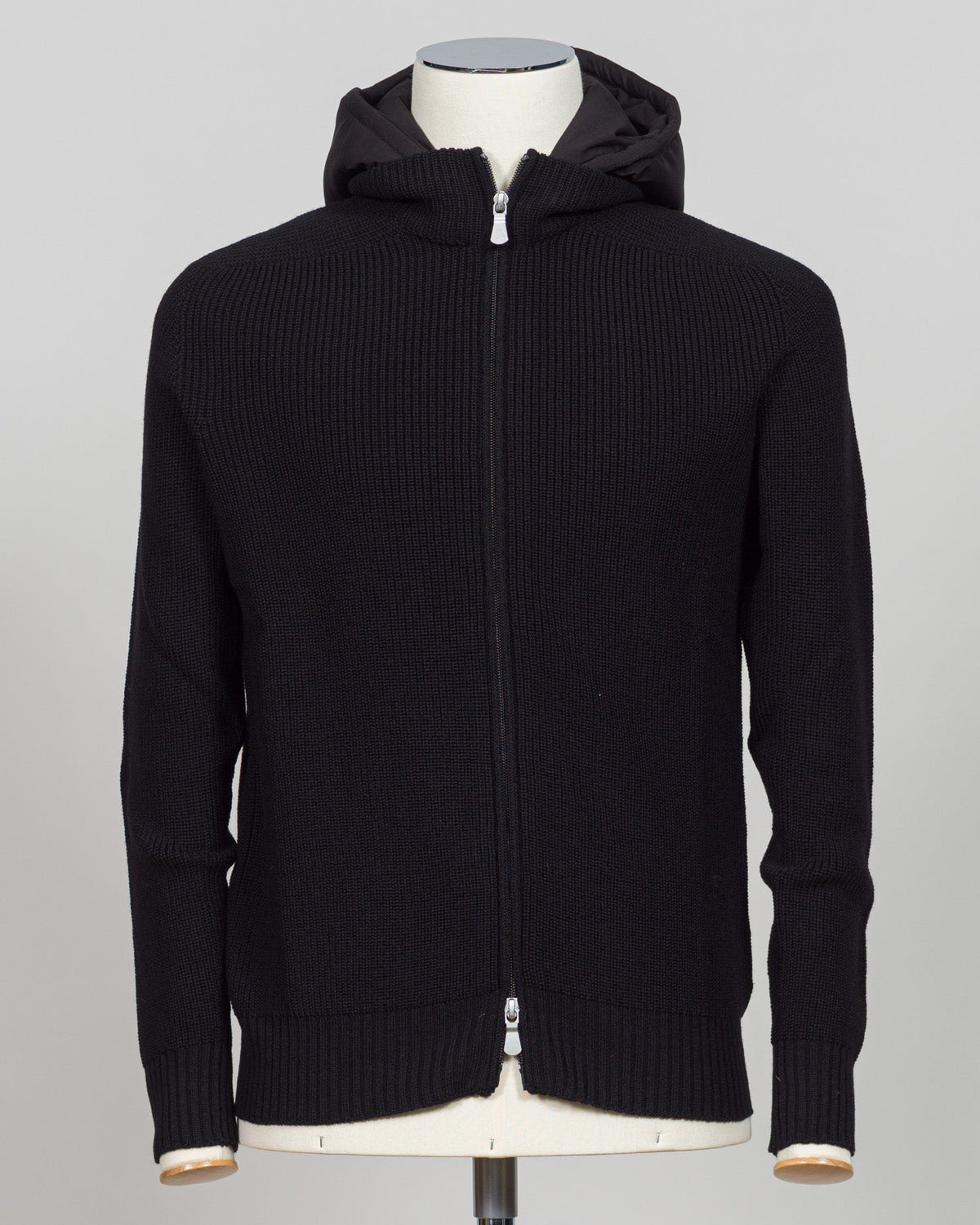 A black ribbed knit zip-up hoodie jacket displayed on a mannequin torso against a light gray background. Italian craftsmanship meets weatherproof functionality in this Gran Sasso rain wool hoodie. Constructed from premium wool blended with water-resistant properties, it delivers warmth and protection without bulk. The relaxed fit and classic black colorway make it a versatile layering essential for unpredictable Nordic weather. Finished with refined detailing befitting Gran Sasso's heritage of fine knitwear