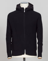 A black ribbed knit zip-up hoodie jacket displayed on a mannequin torso against a light gray background. Italian craftsmanship meets weatherproof functionality in this Gran Sasso rain wool hoodie. Constructed from premium wool blended with water-resistant properties, it delivers warmth and protection without bulk. The relaxed fit and classic black colorway make it a versatile layering essential for unpredictable Nordic weather. Finished with refined detailing befitting Gran Sasso's heritage of fine knitwear