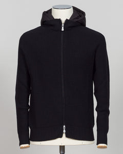 A black ribbed knit zip-up hoodie jacket displayed on a mannequin torso against a light gray background. Italian craftsmanship meets weatherproof functionality in this Gran Sasso rain wool hoodie. Constructed from premium wool blended with water-resistant properties, it delivers warmth and protection without bulk. The relaxed fit and classic black colorway make it a versatile layering essential for unpredictable Nordic weather. Finished with refined detailing befitting Gran Sasso's heritage of fine knitwear