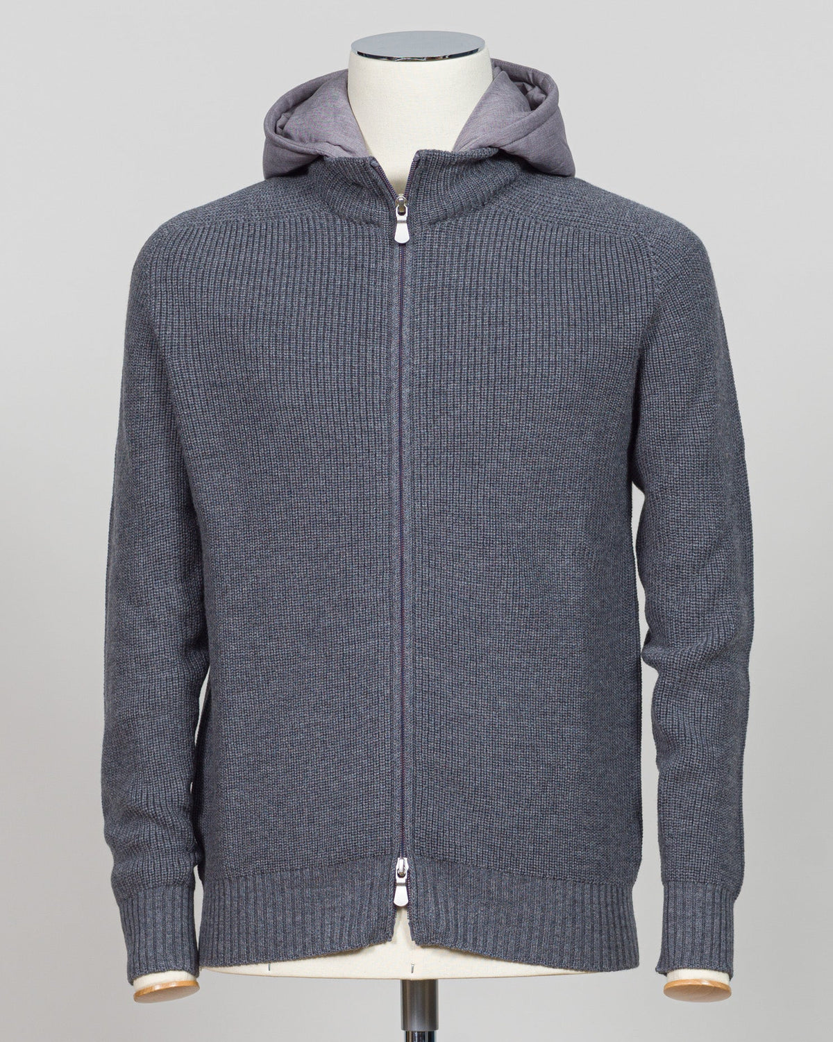 A dark gray ribbed knit zip-up hoodie with a lighter gray hood lining, displayed on a mannequin torso against a neutral light gray Water-repellent merino wool hoodie in warm grey. Engineered for layering and everyday wear, this Gran Sasso piece combines breathable warmth with practical weather resistance. Perfect for transitional seasons and active lifestyles. Gran Sasso Rain Wool Hoodie / Greybackground.