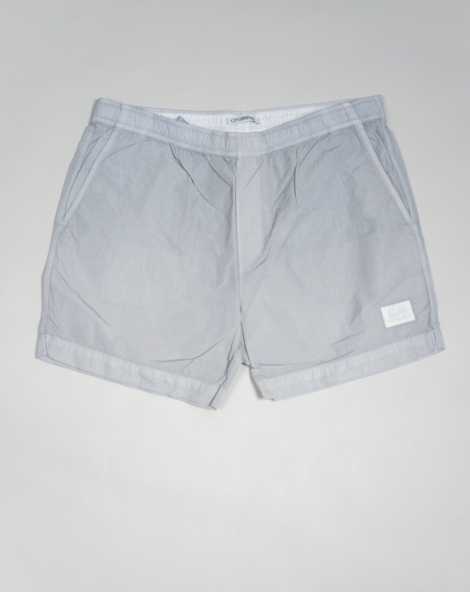 Eco-Chrome swim shorts constructed with an elasticated drawstring waist, hand pockets and a logo detail. Chrome is C.P. Company’s foundation woven multifilament nylon. This new version uses Econyl® regenerated nylon yarns, created through the upcycling of fishing nets.  Art.  BW005A 6369G Col.  936 Light Gray Elasticated drawstring waist Hand pockets Logo detail Nylon dyed 