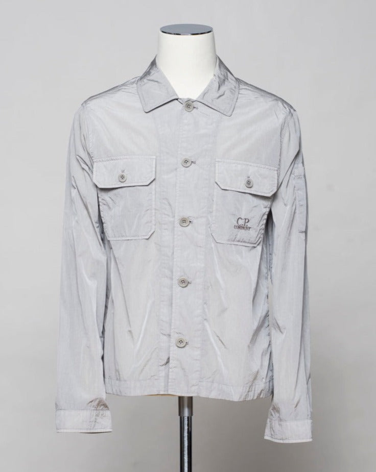Button fastening in front. 2 breast pockets with buttons. Small pocket on left arm with snap fastener. Embroidered tone-on-tone C.P. Company logo on left pocket. Article: 16CMOS106A 005904G. Composition: 100% pre-consumer recycled nylon. Garment dyed. Color: 913 / Drizzle Grey