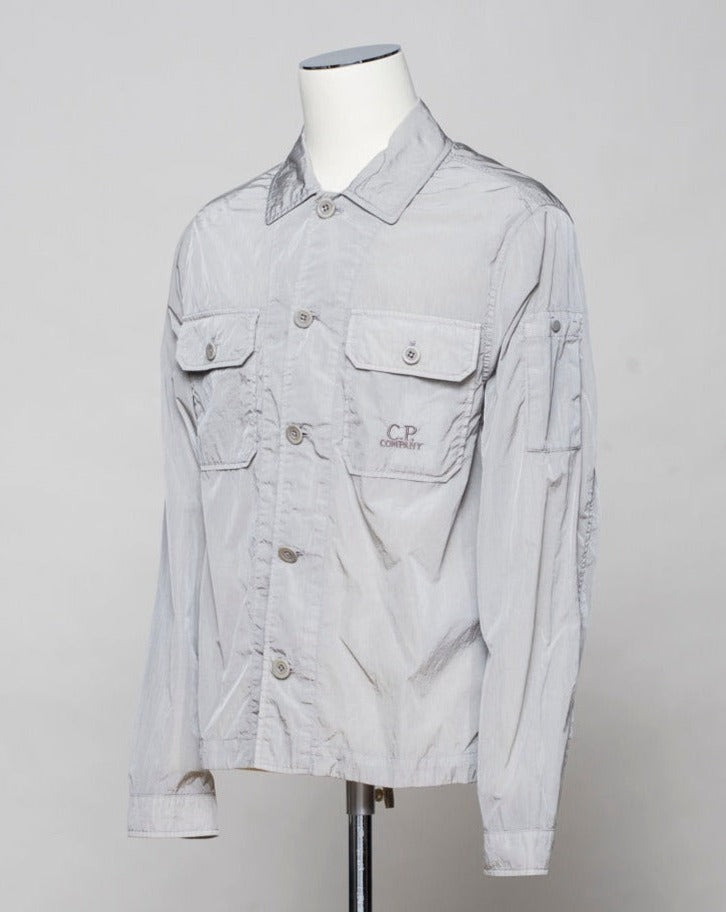 Button fastening in front. 2 breast pockets with buttons. Small pocket on left arm with snap fastener. Embroidered tone-on-tone C.P. Company logo on left pocket. Article: 16CMOS106A 005904G. Composition: 100% pre-consumer recycled nylon. Garment dyed. Color: 913 / Drizzle Grey