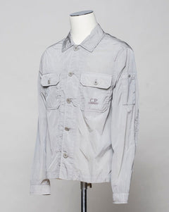 Button fastening in front. 2 breast pockets with buttons. Small pocket on left arm with snap fastener. Embroidered tone-on-tone C.P. Company logo on left pocket. Article: 16CMOS106A 005904G. Composition: 100% pre-consumer recycled nylon. Garment dyed. Color: 913 / Drizzle Grey