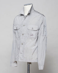 Button fastening in front. 2 breast pockets with buttons. Small pocket on left arm with snap fastener. Embroidered tone-on-tone C.P. Company logo on left pocket. Article: 16CMOS106A 005904G. Composition: 100% pre-consumer recycled nylon. Garment dyed. Color: 913 / Drizzle Grey