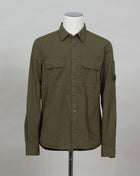 A buttoned shirt featuring a classic collar, secure pockets at the chest, and a discreet sleeve compartment finished with the C.P. Company Lens detail.  Classic Collar Full Button Fastening Secure Chest Pockets Lens Detail Sleeve Pocket Col. 683 / Ivy Green 100% Cotton Art. 12CMSH088A 002824G