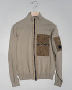 Cardigan with a stand collar, full zip fastening and secure chest compartments. The garment is completed with ribbed hem and cuffs and a C.P. Company Lens detail on the sleeve pocket. Crafted in premium cotton, with piece dyed nylon inserts.  Art. 14CMKN184A 5367M Col. 339 Sand Stand collar Secure front compartments Ribbed hem and cuffs Lens detail Full zip fastening