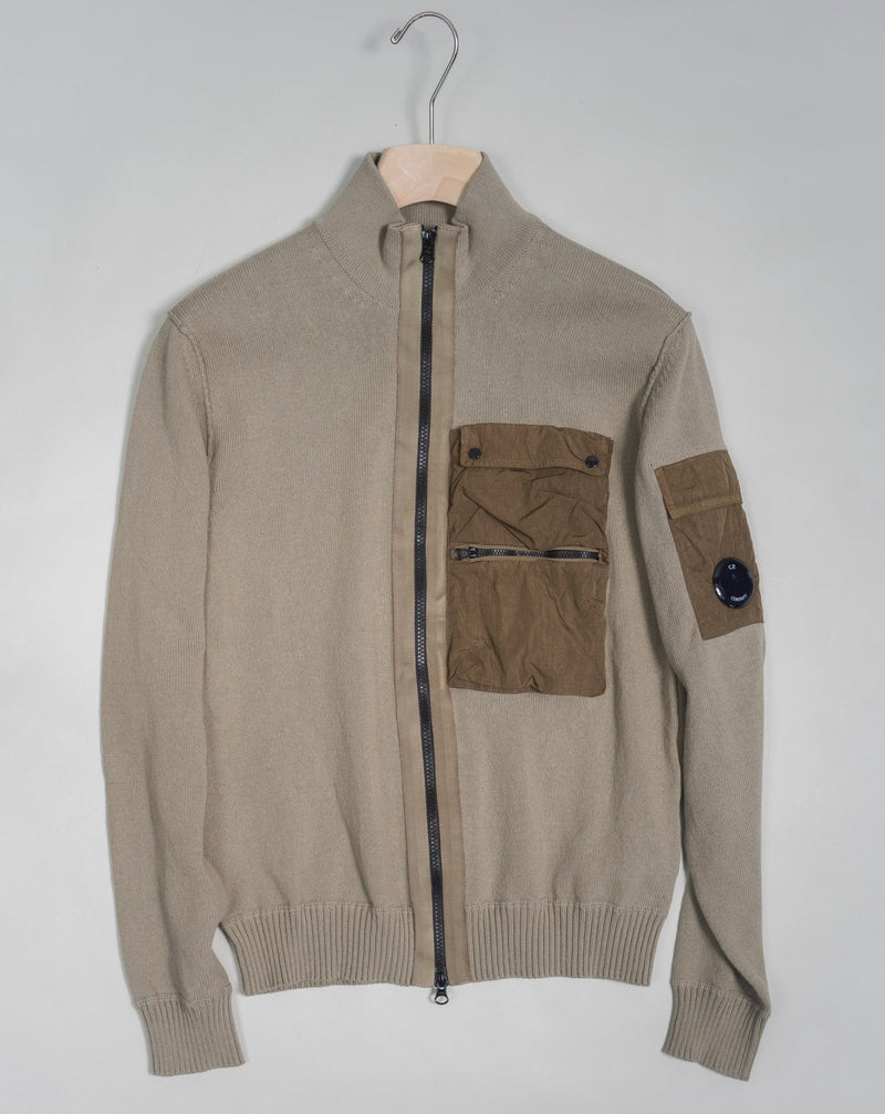 Cardigan with a stand collar, full zip fastening and secure chest compartments. The garment is completed with ribbed hem and cuffs and a C.P. Company Lens detail on the sleeve pocket. Crafted in premium cotton, with piece dyed nylon inserts.  Art. 14CMKN184A 5367M Col. 339 Sand Stand collar Secure front compartments Ribbed hem and cuffs Lens detail Full zip fastening