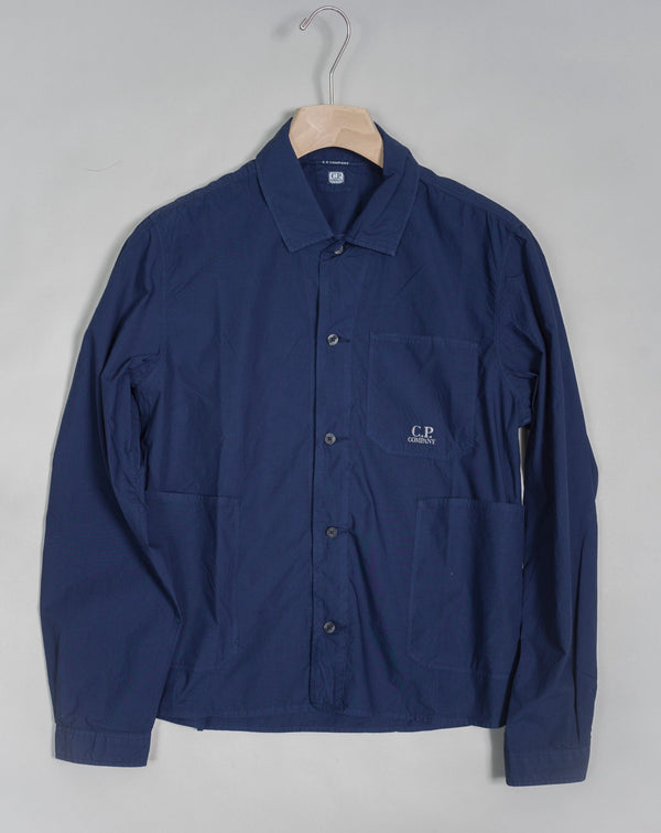 Workwear-inspired long sleeved shirt cut with a classic collar and a full button fastening. The shirt also features an open chest pocket and two open front pockets, adjustable cuffs and a C.P. Company logo detail. Crafted in a crisp cotton poplin with a soft, silken surface, and garment dyed for an increased chromatic depth and intensity throughout. Art. 14CMSH355A 5328G Col. 868 French Blue Cotton Popeline